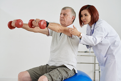 Physioterapist controlling how aged man exercising with dumbbells