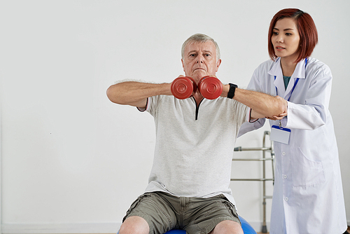 Aged man trying hard to do exercise with dumbbells properly under control of physical therapist