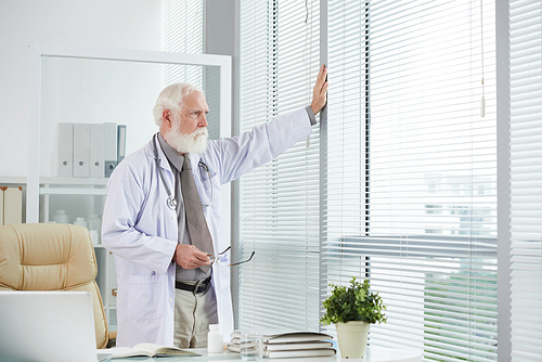 Pensive senior physician standing by window in his office