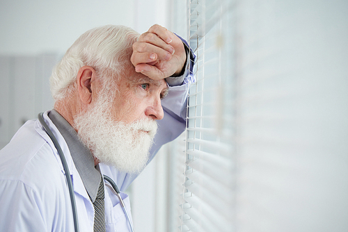 Thoughtful aged general practitioner looking through window in his office