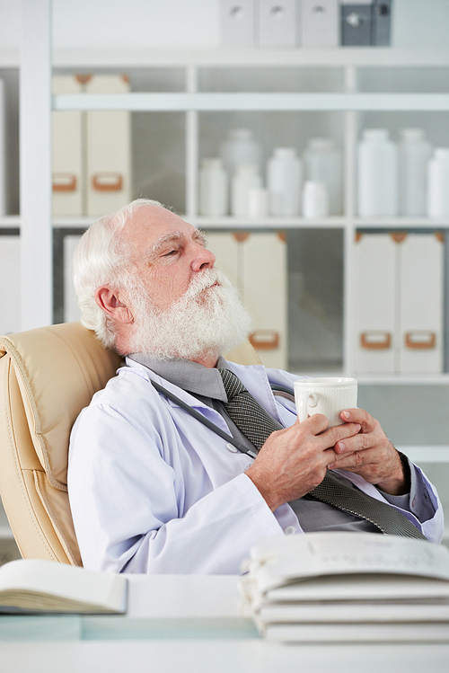Tired elderly doctor relaxing over cup of tea in his office