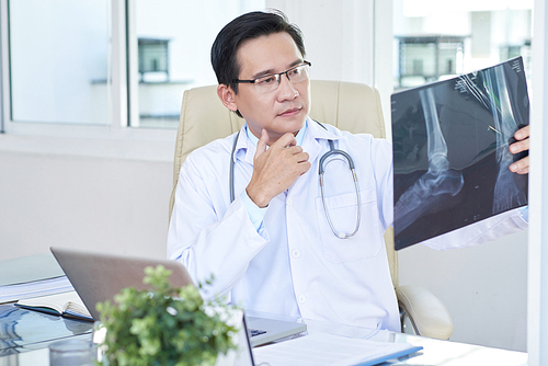 Serious male medic sitting in white coat and looking at X-ray image