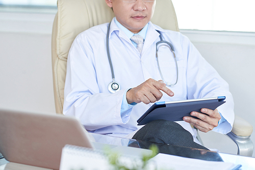 Experienced doctor sitting in his armchair and working in tablet computer