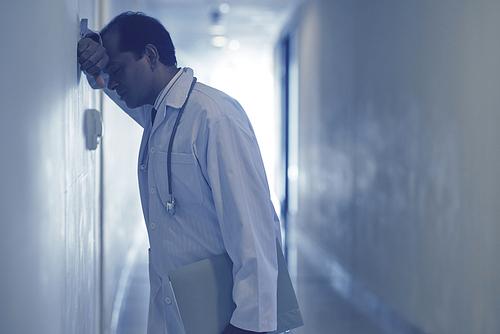 Upset male doctor standing at corridor and leaning against the wall