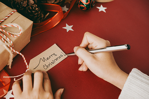Close-up of unrecognizable woman sitting at table with red cloth and writing Christmas wish on tag while preparing Christmas presents