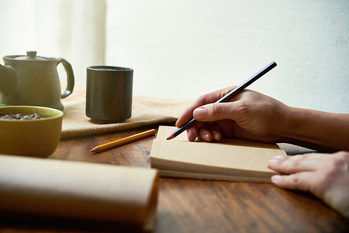 Close-up of female hands holding pencil and writing daily routine in her notebook at the table with cup of tea