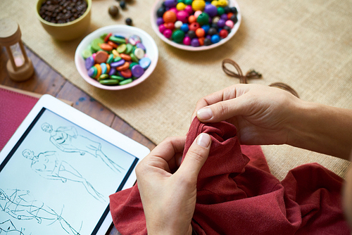 Close-up of female tailor decorating dress with beads using her sketches on tablet pc, handmade at the table