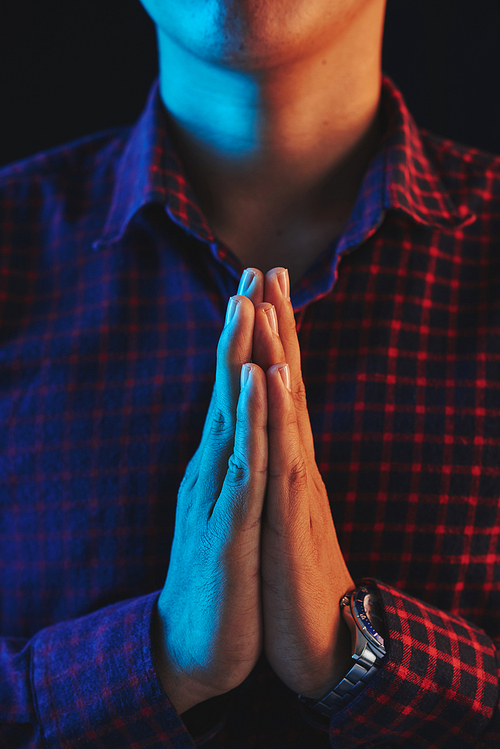 Crop male in checkered shirt and watch on hand holding palms praying on black background