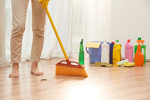 Unrecognizable barefoot woman sweeping floor with broom while wrapped up in housecleaning, detergent bottles, sponges and rags located in corner of room