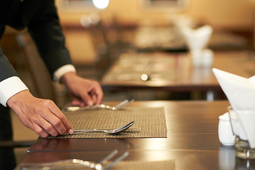 Crop elegant hotel waiter arranging silverware on napkin on table in restaurant