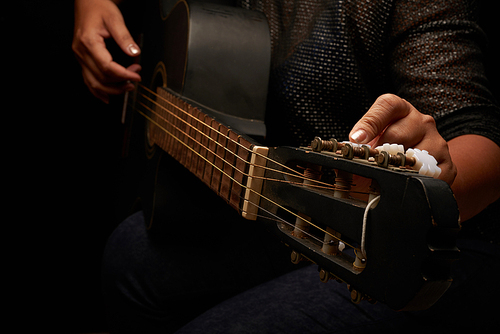 Unrecognizable woman wearing jeans and blouse adjusting strings of acoustic guitar, black background