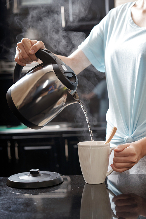 Crop woman holding kettle and filling mug with hot water while brewing drink in modern kitchen
