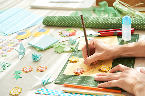 Close-up shot of unrecognizable woman wrapped up in creation handmade greeting card for her mother while sitting at home, messy table surface