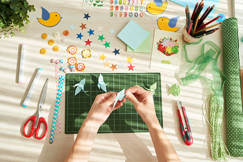 Directly above view of talented woman making origami cranes while sitting at wooden table, its surface covered with colorful decorative items