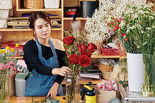 Young pretty Asian florist working at counter and putting fresh flowers into vase