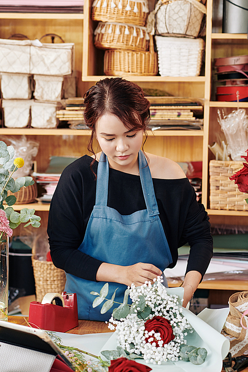 Serious young woman working at her studio and wrapping bouquet in pastel color paper