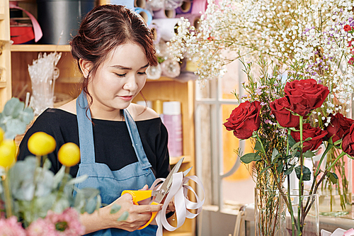 Pretty young Asian florist cutting ribbon to decorate bouquet of flowers