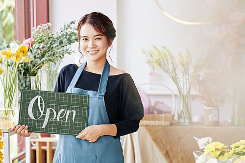 Pretty young female flower shop owner holding open sign and smiling at camera