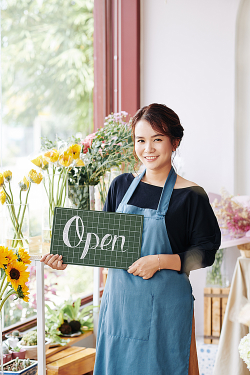 Lovely smiling Vietnamese florist in blue apron holding open sign and 
