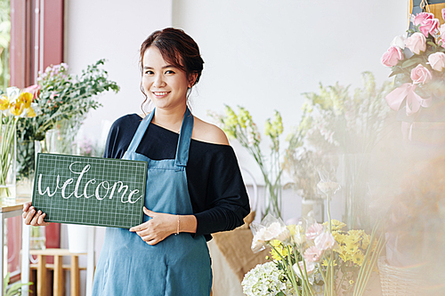 Pretty young Vietnamese florist holding welcome signboard and welcoming customers to her flower shop