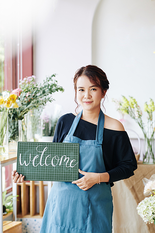 Portrait of young Asian woman in aprons holding welcome signboard and inviting customer to her flower shop