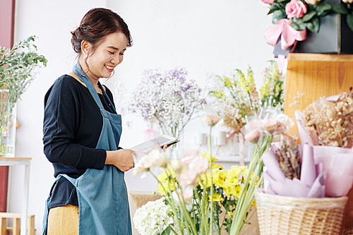 Pretty smiling young florist using tablet computer when checking flowers in her store