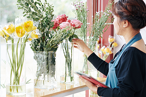 Professional florist checking order on tablet computer and choosing flowers from vases on shop window sill