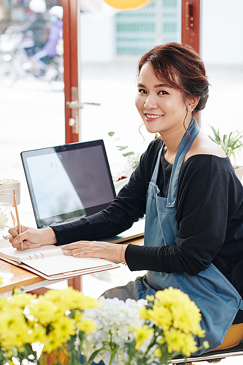 Portrait of young positive flower shop owner in apron sitting at her desk and writing in planner