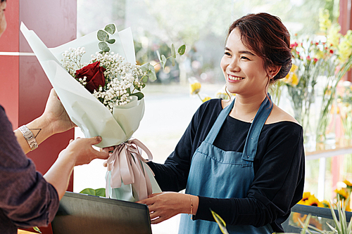 Positive flower shop worker giving beautiful bouquet with red rose to customer