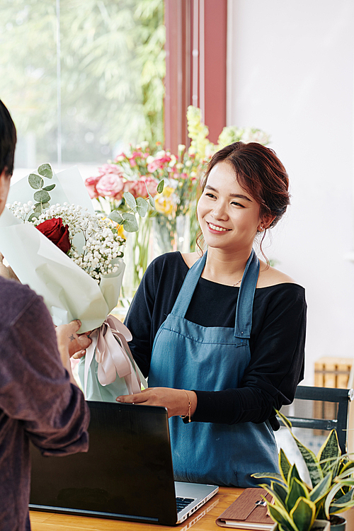 Cheerful pretty florist in blue apron giving bouquet of flowers to customer