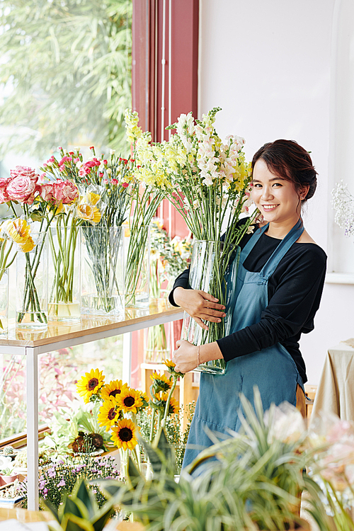 Cheerful young Asian florist standing among flowers with transparent glass vase