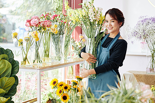 Smiling young Asian woman placing vases with various beautiful blooming flowers in flower boutique