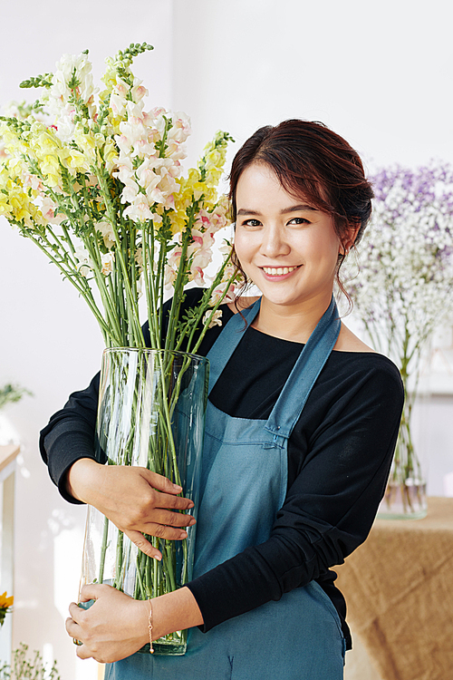 Happy young flower shop owner holding big glass vase with tall blooming flowers