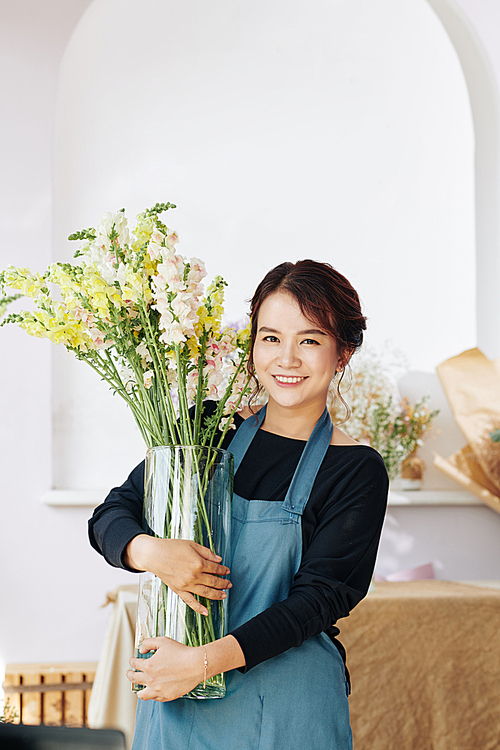 Portrait of young happy woman in blue apron holding big glass vase with blooming flowers