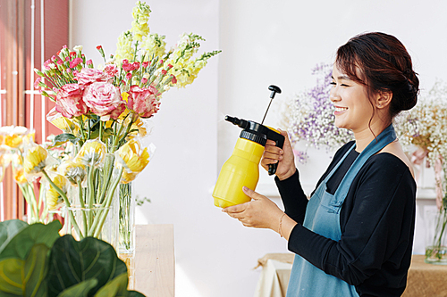 Cheerful young flower shop worker spaying fresh flowers in glass vases
