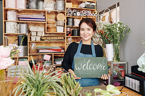 Portrait of happy young florist standing at counter in slower shop with welcome signboard