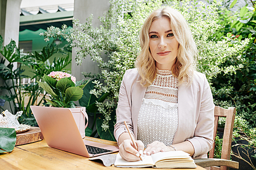 Portrait of beautiful smiling business lady sitting at outdoor table with opened laptop and taking notes in planner