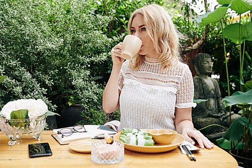 Beautiful elegant young woman drinking cup of coffee and eating spring rolls for business lunch in outdoor cafe