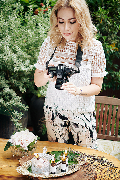 Pretty female photographer checking photos of organic cosmetics she made with professional digital camera