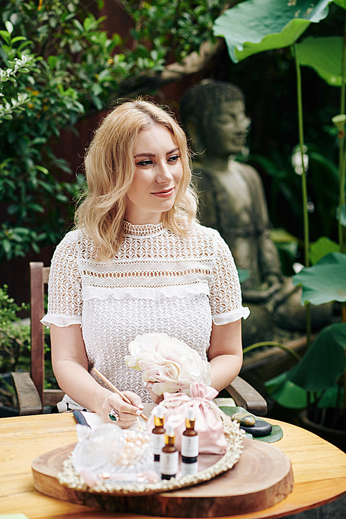 Smiling beautiful young woman sitting at table in backyard and writing business plan for her cosmetics brand