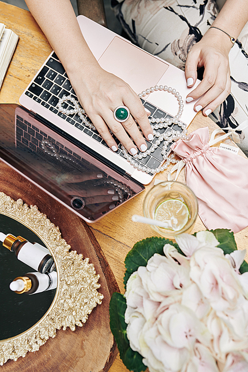 Hand of woman wearing ring with serpentine stone and putting hand on pearl beads on opened laptop at her vanity