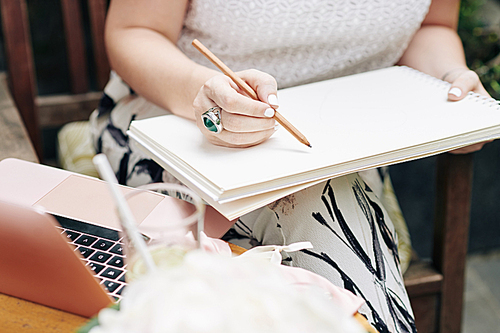 Close-up image of businesswoman drawing logotype or sketch with pencil in her sketchbook when sitting outdoors at table