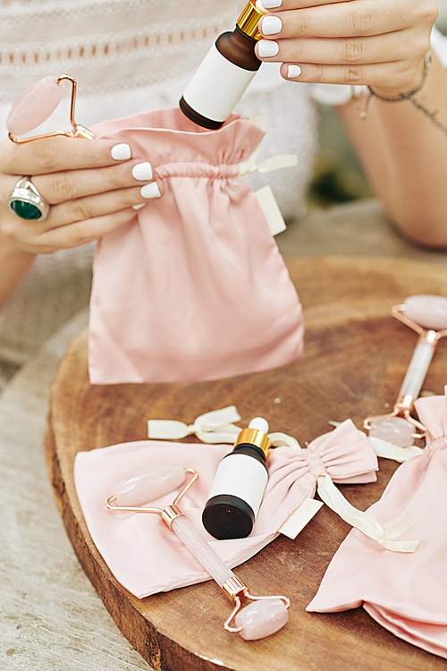 Close-up image of small cosmetics company owner packing set of rejuvenating essence and quartz massage roller into silk pink sacks