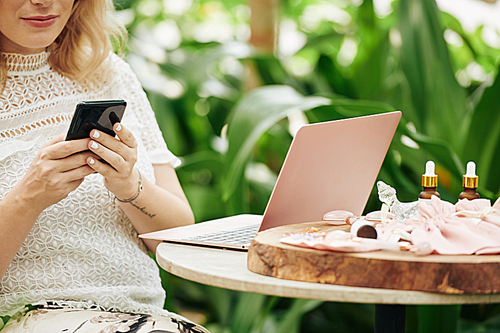 Cropped image of young woman sitting at cafe table with laptop and beauty products and answering questing from client on social media