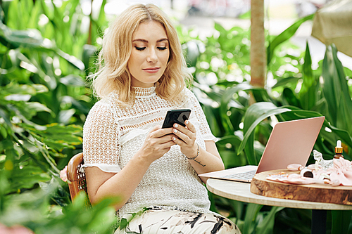 Beautiful young woman sitting at table in outdoor cafe among lush green foliage and reading artlicle on smartphone