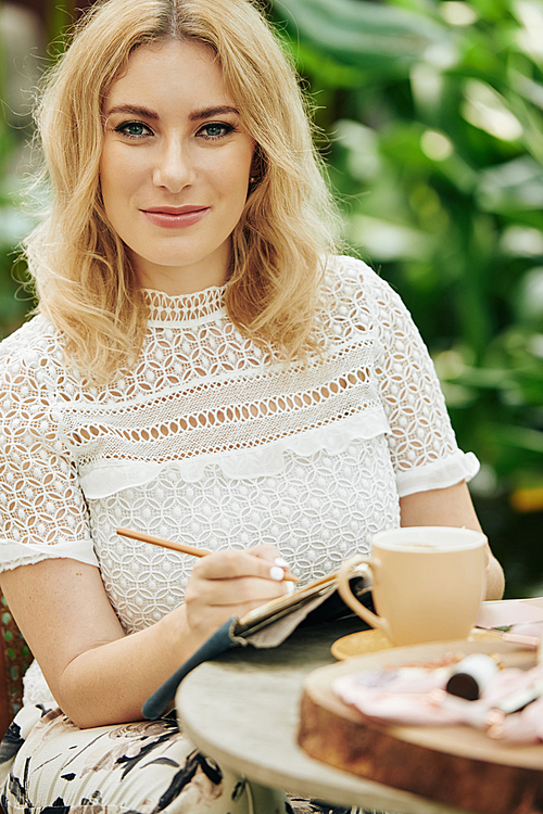 Smiling elegant young blond woman sitting at cafe table, drinking mug of coffee and writing in planner