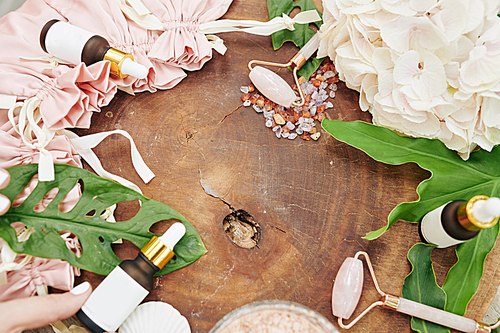 Wood stump with face massage rollers, serums, flowers and leaves, view from above