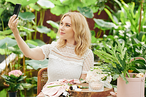 Attractive smiling young blond woman sitting outdoors with natural cosmetics on table in front of her and taking selfie