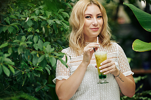 Beautiful smiling woman enjoying tasty sour lemon lime alcoholic cocktail when walking in garden