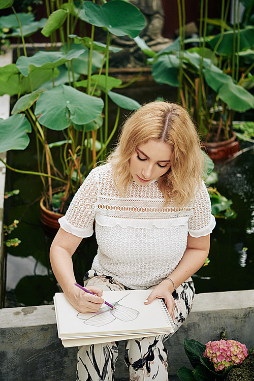 Creative young woman sitting at small garder pond and drawing water lily leaves